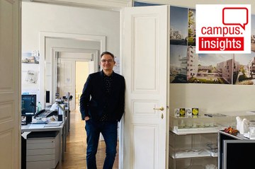 Architekt Sascha Bradic lehnt im Architektur-Büro in einem klassischen Altbau mit weißen Flügeltüren an einem Türrahmen und lächelt in die Kamera - im Raum sieht man Bilder und Modelle von Bauprojekten